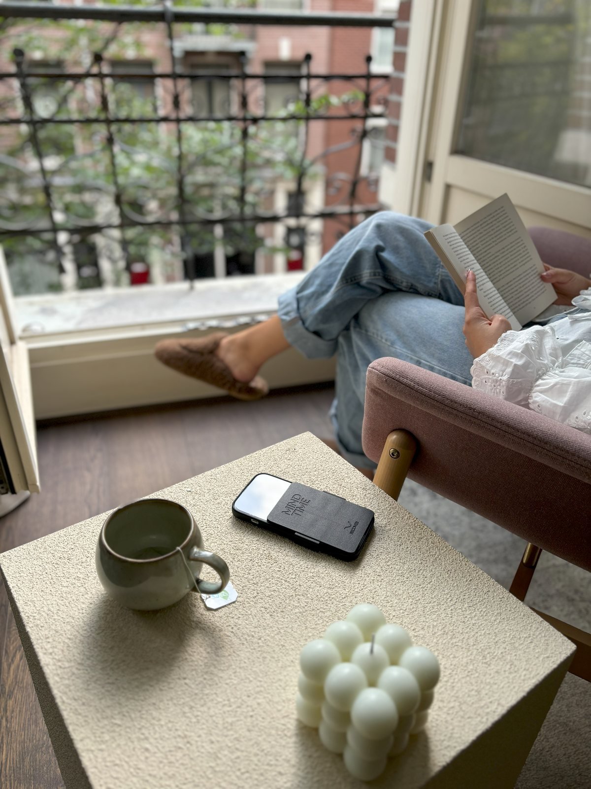 Reading in an armchair by an open balcony door; the phone with ReCover rests on the side table next to a cup of tea